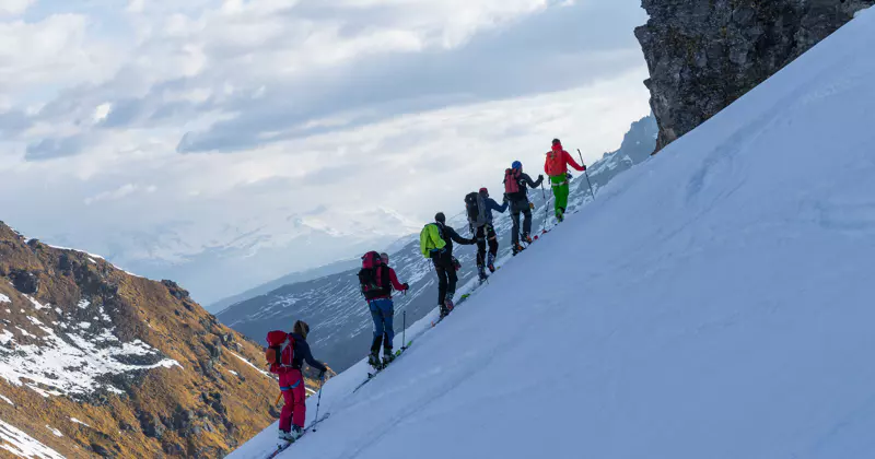 Een groep skiërs in kleurrijke uitrusting beklimt in een rij achter elkaar een steile, besneeuwde berghelling, met rotsachtige kliffen en besneeuwde bergtoppen in de verte op de achtergrond, onder een gedeeltelijk bewolkte hemel.